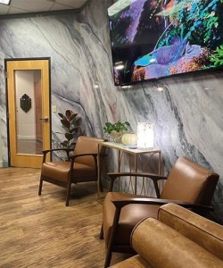 Modern dental office waiting room featuring leather armchairs, marble-patterned walls, and a TV screen.